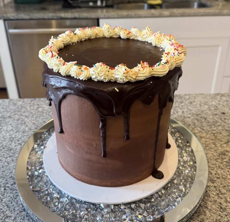 Tall chocolate cake with a dark chocolate drip and a piped cream border with rainbow sprinkles