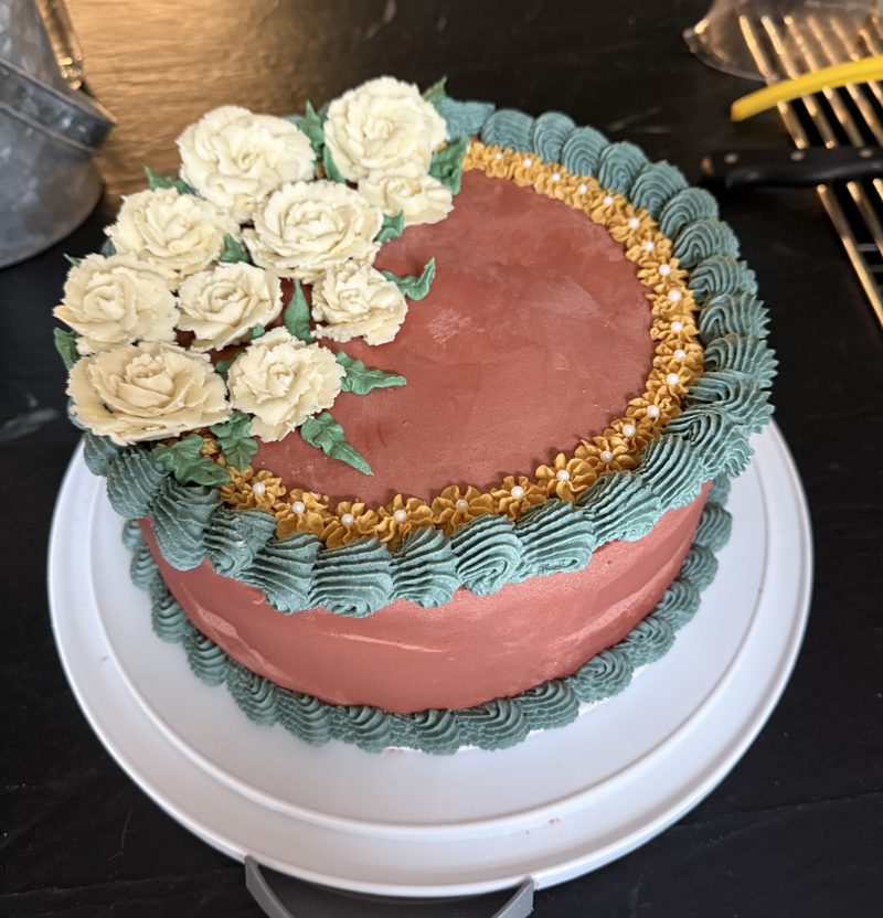 Terracotta buttercream cake with piped cream flowers, a sage-green shell border and a gold ruffle set with sugar pearls