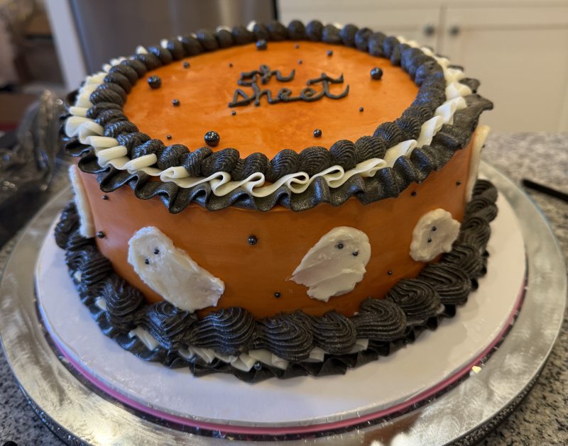 Orange Halloween cake reading &ldquo;oh sheet&rdquo;, with buttercream ghosts, black and white ruffles and black sugar pearls